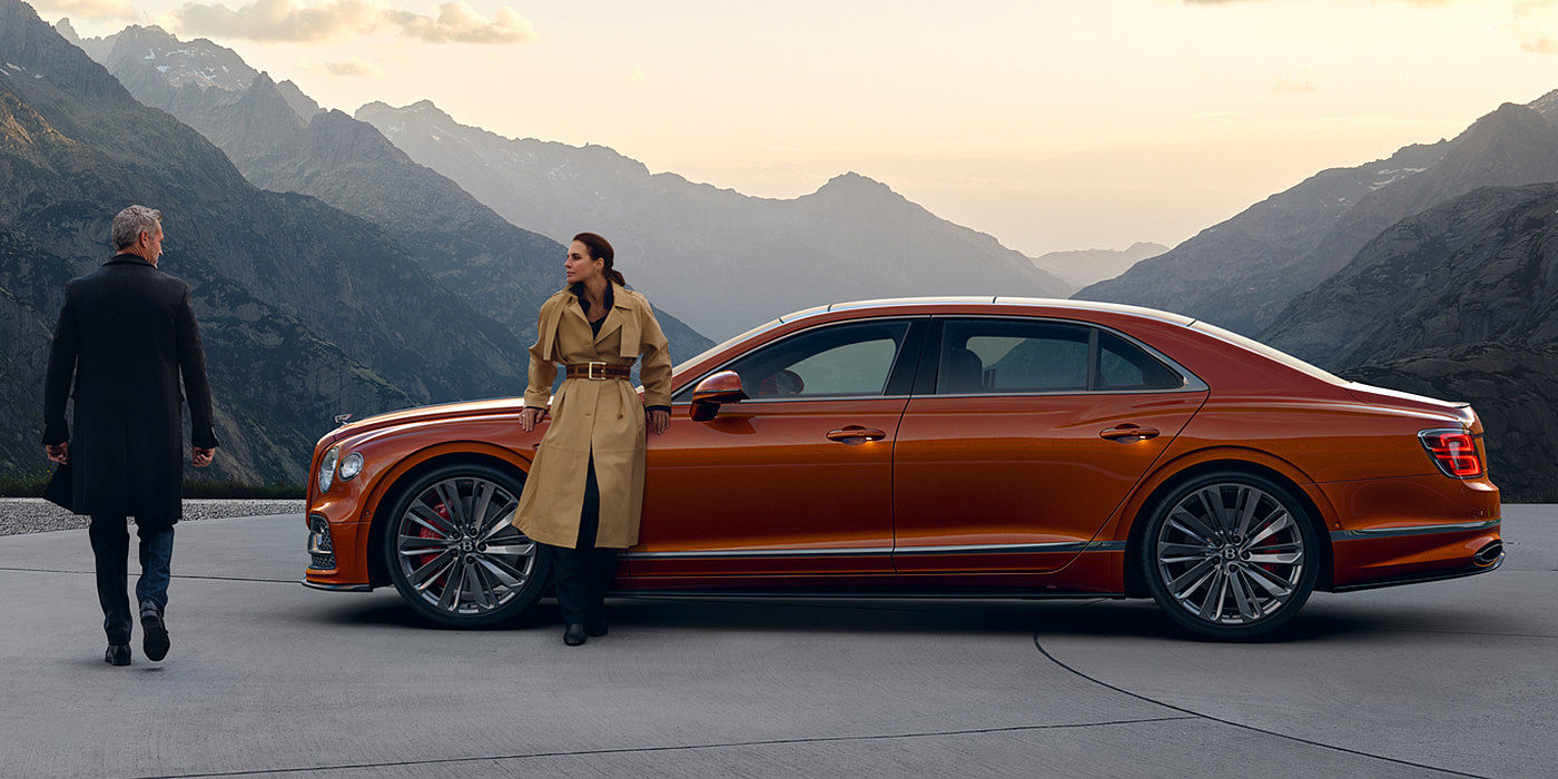 Bentley Antwerp Bentley Flying Spur Speed parked in Orange Flame coloured exterior parked, with mountainous background and two people in view.